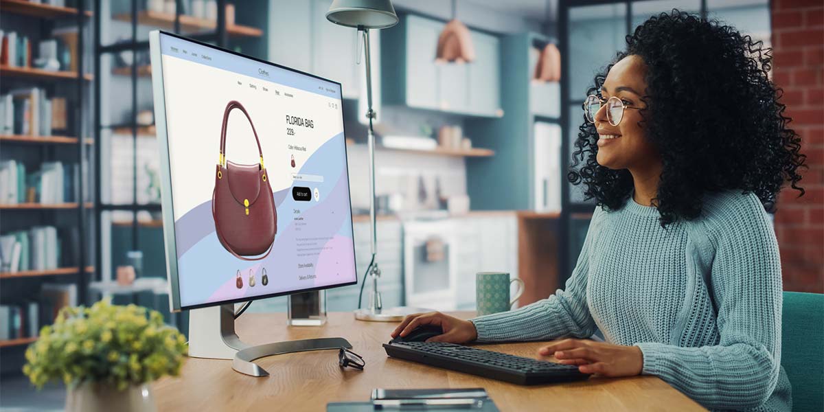 woman with curly black hair and glasses sitting at a desk and looking at a bag on an ecommerce site