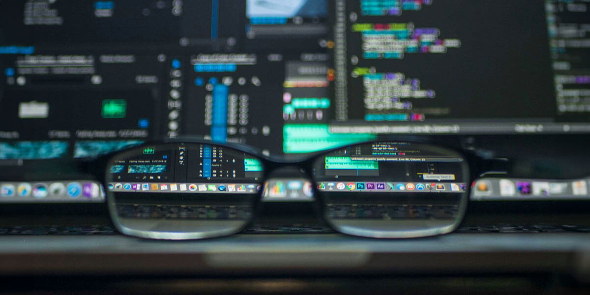 A pair of eyeglasses rests on a laptop keyboard, reflecting lines of code and cybersecurity tools on the screen.