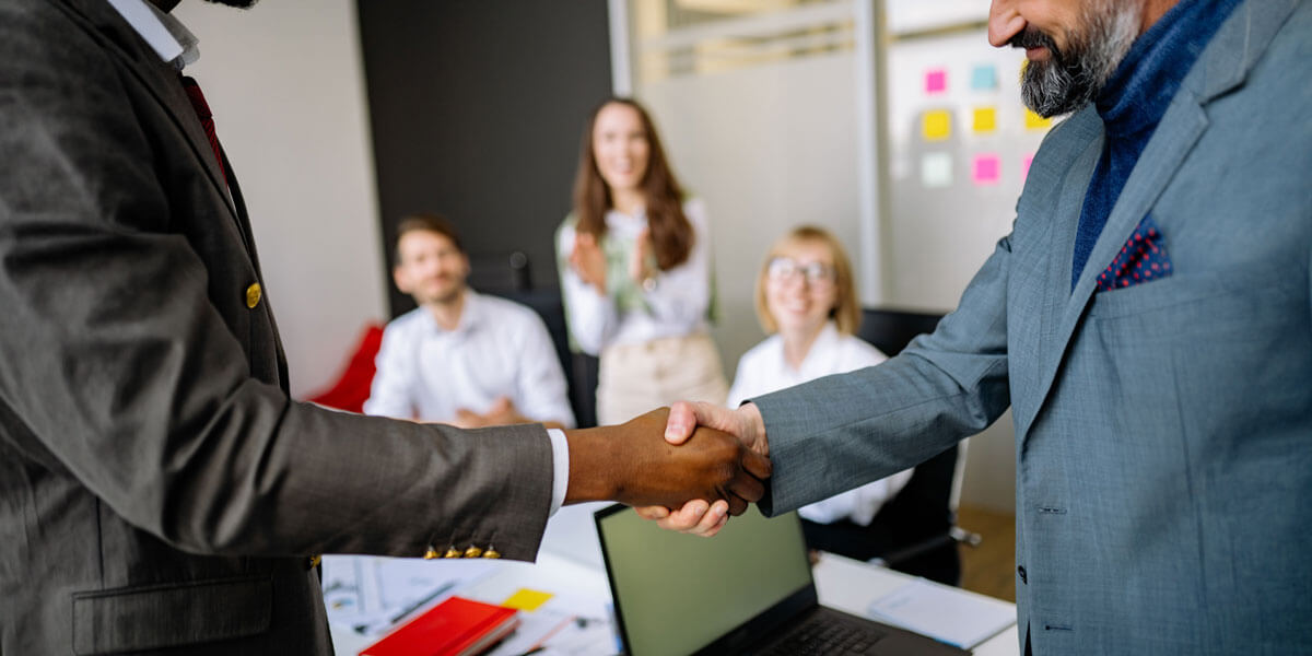 Two businessmen shaking hands while colleagues applaud in the background of an office.