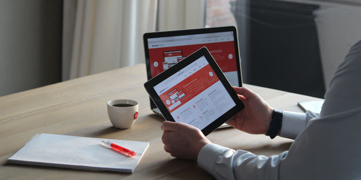 Person reviewing a website design on a tablet with a laptop and coffee cup on the desk.