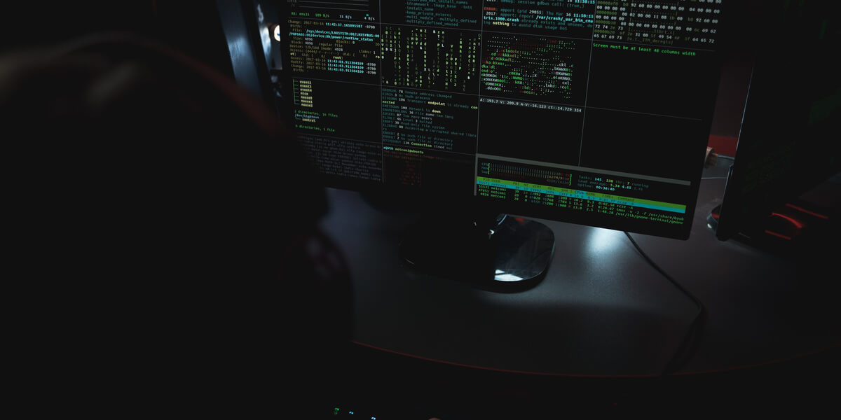 A person sits at a computer with code displayed across multiple monitors in a dark room.